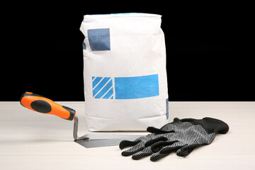 Bag of cement powder, putty knife and rubber gloves on light wooden table against black background....