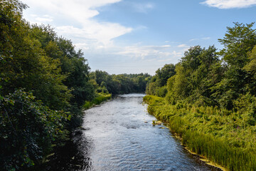 A river in a deciduous forest on a sunny summer evening. One bank in the shade, the other in the sunlight. Blue sky with clouds and cloudy haze. The riverbed is crossed by high-voltage power lines