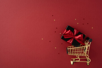 A golden shopping cart carries a beautifully wrapped gift box against a festive red background, symbolizing Black Friday present shopping