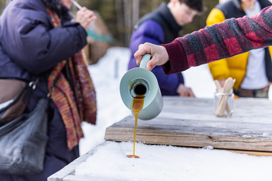 Person is pouring maple syrup onto fresh snow to make maple taffy candy on a rustic wooden table outside in winter. Selective focus