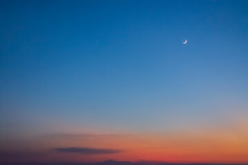 A wide angle shot of a blue-orange sky with the horizon disappearing and a crescent moon in the distance.