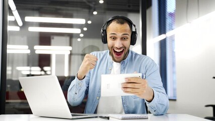Excited african american businessman in wireless headphones watching sports match or competition using digital tablet sitting at workplace in business office. Happy man cheering for favorite team - Powered by Adobe