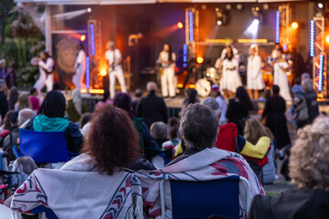 Couple is watching a concert on an outdoor stage at dusk, sitting on camping chairs and covered with a blanket. Selective focus