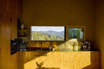 A scenic view of lush green mountains through an open kitchen window. The golden light illuminates the landscape, blending the natural beauty outside with the warm tones inside