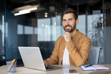 Young professional confidently engaged with laptop in modern office. Positive expression indicates focus and productivity. Smartphone, stationery enhance workspace, providing efficient
