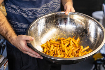 Chef is holding a large stainless steel bowl of freshly cooked french fries sprinkled with spices. Selective focus