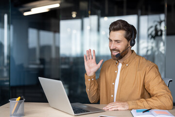 Smiling man in brown shirt using laptop headset for video call in modern office setting. Engaging in virtual meeting with colleagues, demonstrating innovation communication technology.