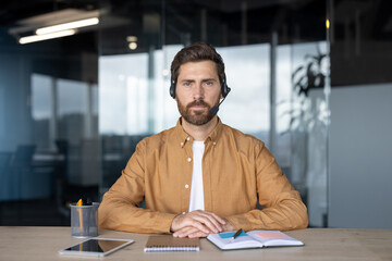 Professional man with headset at office desk using tablet for communication. Focused environment with modern workspace essentials like notebook and pen, embodying multitasking and digital interaction