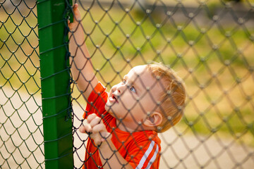 little boy behind iron fence on summer playground, children lifestyle
