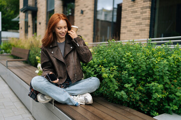 Joyful young redhead female in leather jacket sitting on urban bench, enjoying cup of coffee and using mobile phone, taking break from day to relax and catch up with friends on summer day outdoors.