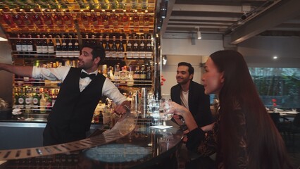 Smart bartender with VIP customer at luxurious bar greeting new beautiful guest at counter in engaging lively conversation at back view in lighting atmosphere at lighting warm nightclub. Vinosity.