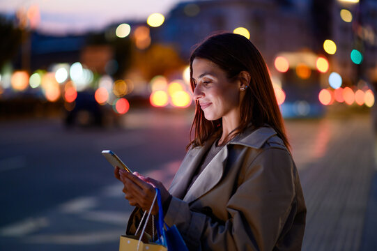 Elegant woman is using smartphone on city street at night - Powered by Adobe