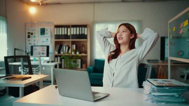 Young asian businesswoman is working on her laptop in her bright office. She takes a break from work, stretching her arms and relaxing