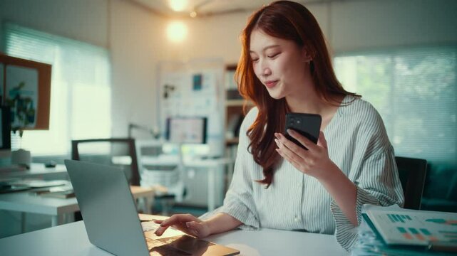 Young asian businesswoman is multitasking at her desk, smiling while using her laptop and smartphone in her modern office