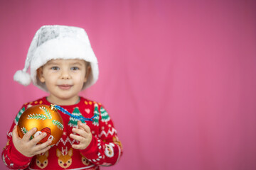 Joyful child in festive sweater and Santa hat posing for Christmas photo. Cute kid holding decoration in hands isolated on pink background. Child looking forward to new year and Christmas.