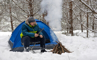 A tourist dressed in winter clothes lights a fire with a lighter near a tent in a snowy forest. A man warms himself by a fire after laying out a tent in a remote wilderness area in winter.