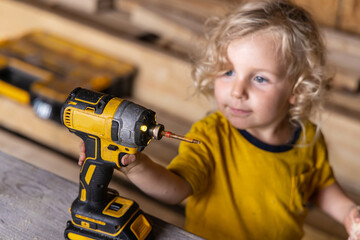 Curious boy with blond hair assembles furniture at home using a cordless drill, showing interest in carpentry and construction. Selective focus
