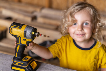 Young boy happily holds a cordless drill in a workshop, showing curiosity and engagement in learning about tools. His blonde hair and bright smile add to the playful scene. Selective focus