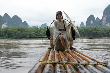 Hanfu girl holding an umbrella poses on bamboo raft on river with two cormorants. Xingping, China