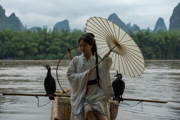 Hanfu girl seated on a bamboo raft holds an umbrella while fixing her hair