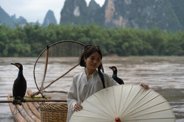 Hanfu girl holding an umbrella poses on bamboo raft on river with two cormorants. Xingping, China