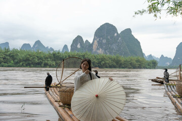 Barefoot girl in Hanfu clothing stands with an umbrella on a bamboo raft, Yangshuo County, China