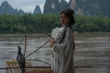 Hanfu girl smiles while seated on a bamboo raft, holding a round fan with cormorant birds beside her