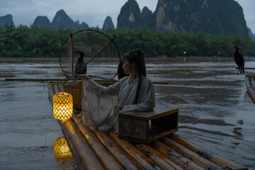 Hanfu girl holding an illuminated lantern on a bamboo raft with Cormorant birds at night