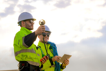 Two engineers wearing high visibility safety jackets and helmets measure wind speed with an anemometer and analyze data on a tablet at a renewable energy site. The cloudy sky adds depth to the scene.