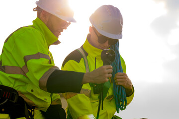 Two engineers wearing high visibility safety jackets and helmets measure wind speed with an...