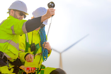 Two engineers wearing high visibility safety jackets and helmets measure wind speed with an...