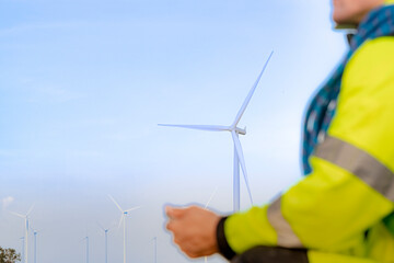 Workers in high visibility jackets examine a wind turbine farm, one pointing towards the turbines while the other holds a mobile device. The scene emphasizes renewable energy and modern technological.