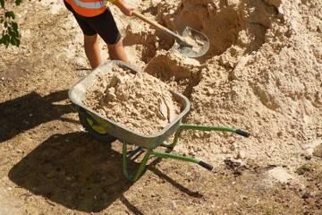 Hands of a worker with a shovel fill a wheelbarrow with sand against a pile of sand