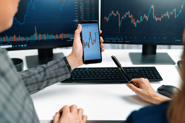 Cropped pointing hand with pen in businessman, checking with holding smartphone by woman investor online website program application with dynamic stock market at home office at back side. Infobahn.