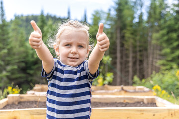 Young boy with long blond hair is giving two thumbs up while standing in a garden. Selective focus