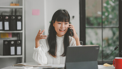 Asian businesswoman is working in her bright office, talking on the phone and gesturing while looking at her laptop