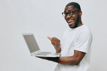 Happy young man in casual white t shirt, smiling and holding laptop, showcasing excitement with thumb up on light background, embodying success and positivity