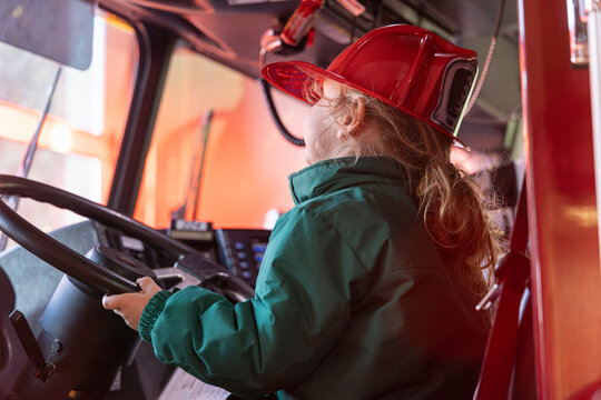 Young boy is sitting in the driver's seat of a firetruck wearing a firefighter's helmet and smiling. Selective focus