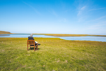 Fototapeta premium Person sits on a wooden chair in a peaceful field by the river, wearing a hoodie, and gazing at the tranquil water and clear blue sky in western Iceland. The scene exudes calm and contemplation