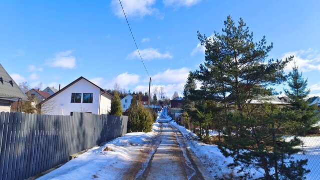 There is snow on the ground. There are wooden and metal mesh fences and residential houses along the street. Ice has formed on the road after the thaw and there is a rut in it. Sunny winter weather