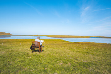 Fototapeta premium Guy sits on a wooden chair in a green field, reading a book while overlooking a peaceful river and clear blue sky in western Iceland. The scene exudes tranquility and quiet reflection