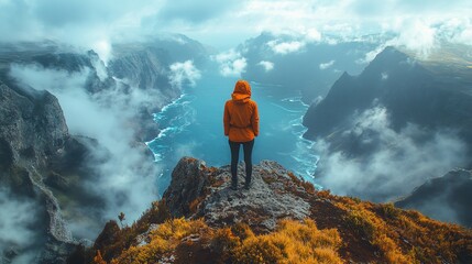 A lone hiker stands on a rocky clifftop overlooking a stunning valley with ocean in the distance, surrounded by clouds and fog.