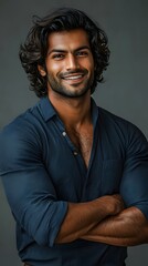 A portrait of an attractive young Indian man with wavy hair, smiling and standing confidently in front of a grey background, wearing a dark blue shirt