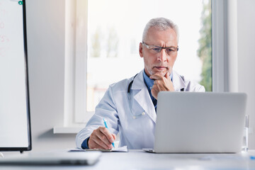 Mature male doctor working on laptop computer and writting notes while sitting in medical office