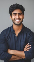 A portrait of an attractive young Indian man with wavy hair, smiling and standing confidently in front of a grey background, wearing a dark blue shirt