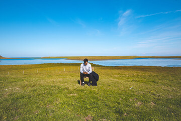 Boy sits on a wooden chair in a green field, looking down at their phone, surrounded by the peaceful scenery of a river and clear blue sky in western Iceland