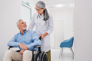 Middle aged woman doctor pushing senior patient in wheelchair in hospital corridor while talking to...