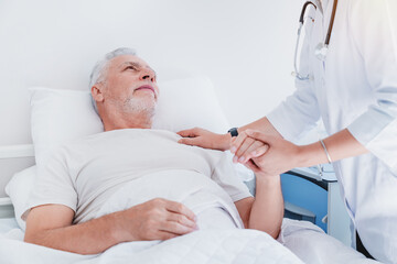 Fototapeta premium Asian female doctor holding hands and supporting sad senior man lying in hospital bed