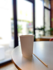Simple white paper cup sits on a round table in a cozy cafe, inviting a moment of relaxation and warmth. Blurred background suggests a peaceful indoor setting, perfect for enjoying a quiet coffee
