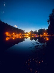 Fototapeta premium pond reflection in the night park, night sky, trees silhouettes and lights reflection on the lake surface